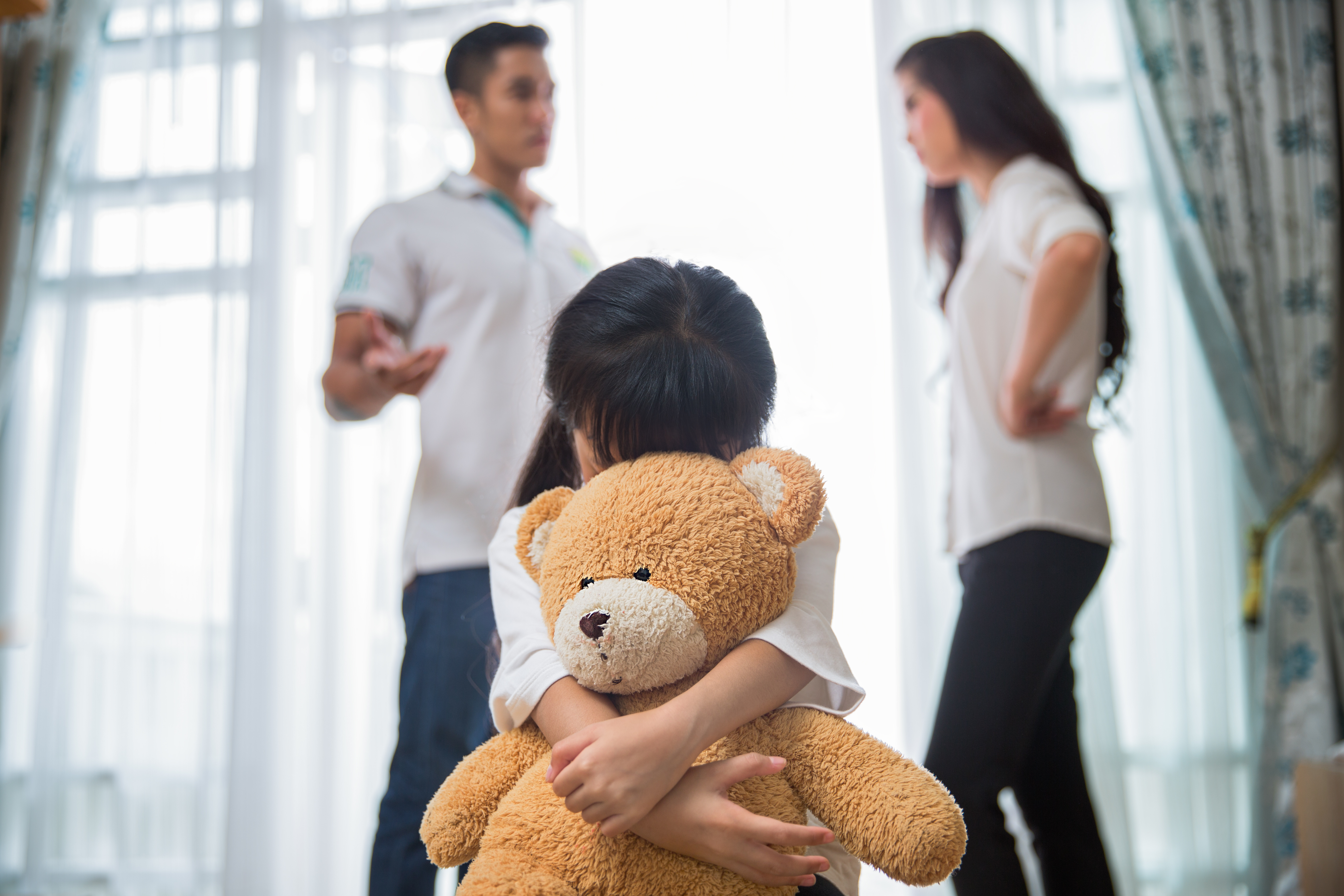 Sad child hugs stuffed bear while father and mother are arguing in the background